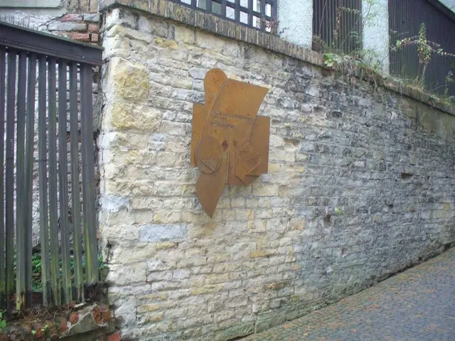 The image shows a stone wall with a metal gate on the left side and a building with windows and...
