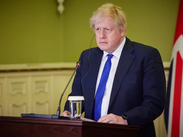The image shows a man in a suit and tie standing at a podium with a microphone, a glass of water,...