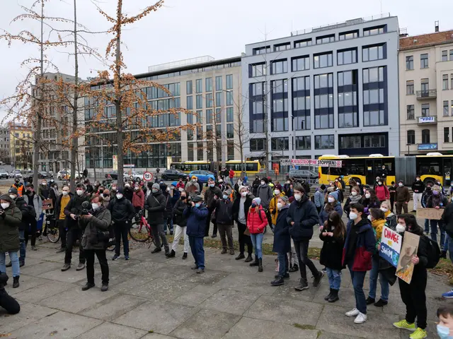 The image shows a large group of people standing in front of a building, some of them wearing masks...