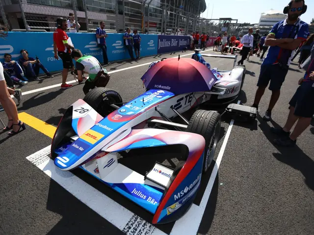 The image shows a group of people standing around a race car on a track, with a stadium in the...