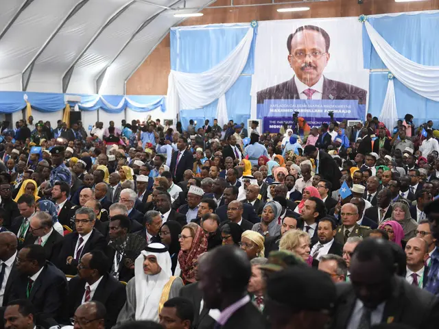 The image shows a large crowd of people standing in front of a banner with a picture of a man on...