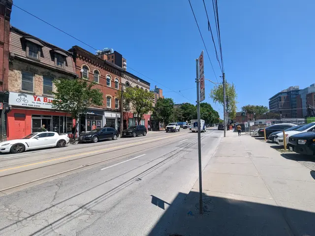 The image shows a city street with cars parked on the side of it, buildings with windows, trees,...
