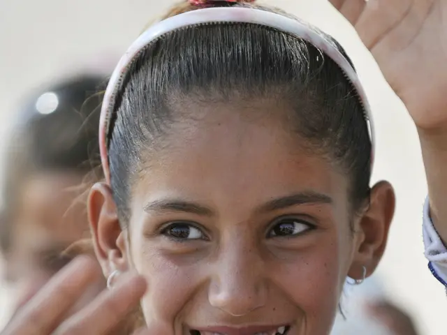 The image shows a young girl in a school uniform smiling and waving to the camera, with a few other...
