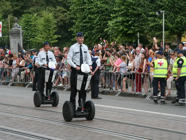 The image shows two police officers riding segways down a street in front of a crowd of people,...