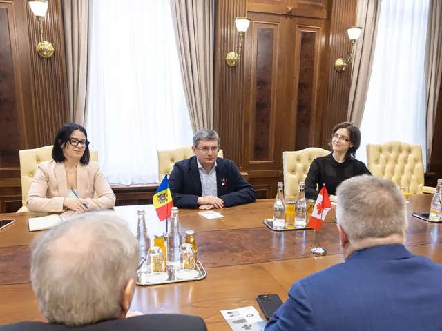 The image shows a group of people sitting around a wooden table with flags, bottles, glasses,...