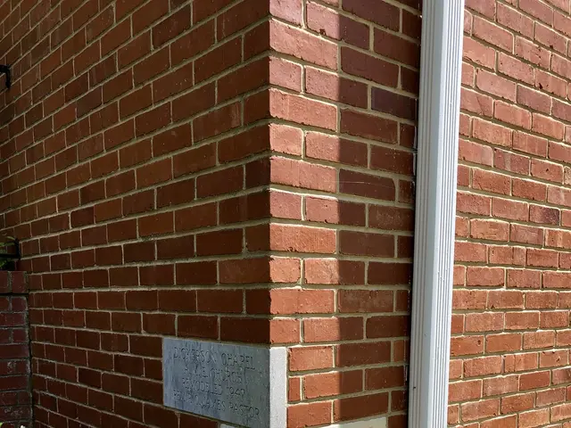 The image shows a brick building with a plaque on the side of it, surrounded by plants on the left...