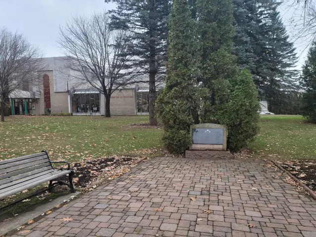 The image shows a park bench sitting in the middle of a brick walkway surrounded by trees, plants,...