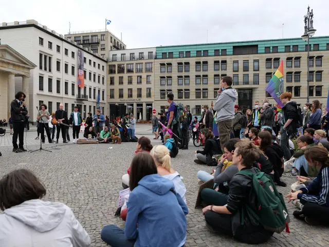 The image shows a group of people sitting on the ground in front of a crowd of people, some of whom...