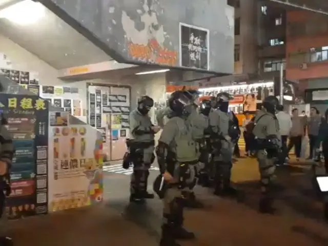 The image shows a group of police officers standing in front of a crowd of people wearing helmets...