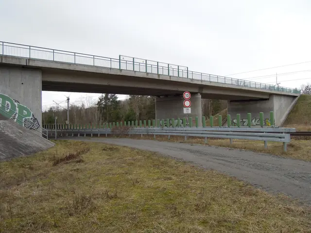 The image shows a bridge over a dirt road with graffiti on it, railings, poles, sign boards, a...