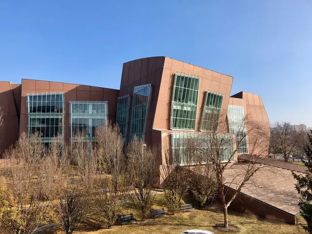 The image shows a large building with a lot of windows on the side of it, surrounded by trees,...