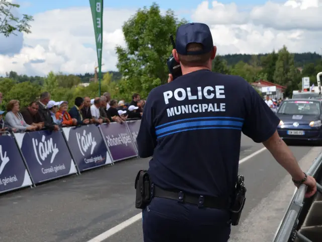 The image shows a police officer standing in front of a crowd of people. He is wearing a cap and...