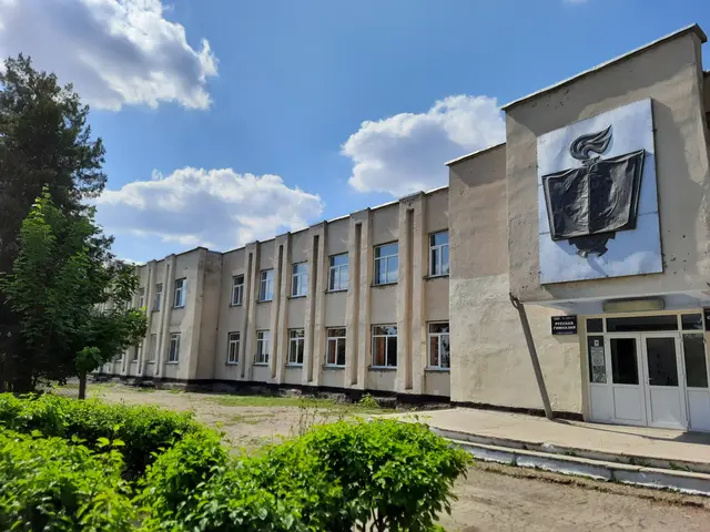 The image shows a large building with a sign on the side of it, surrounded by plants, trees, and a...