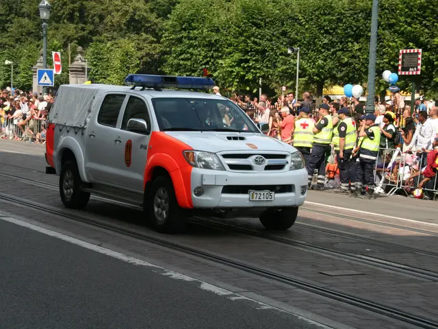 The image shows a police car driving down a street next to a crowd of people, some of whom are...
