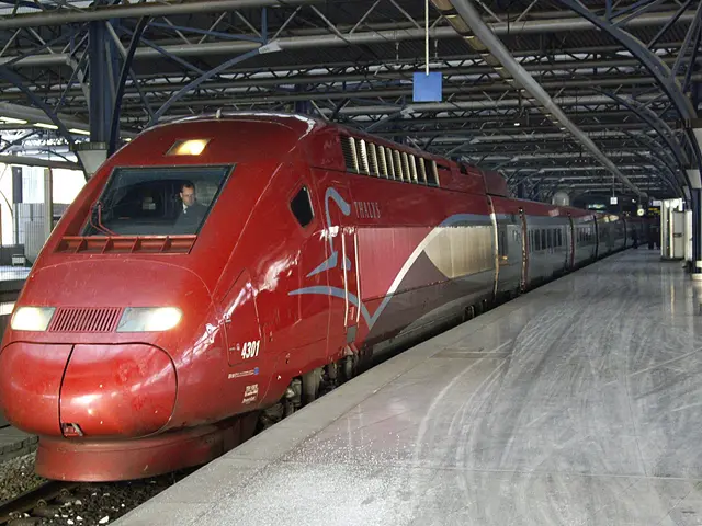 The image shows a red high-speed train parked at a train station, with a platform on the right side...