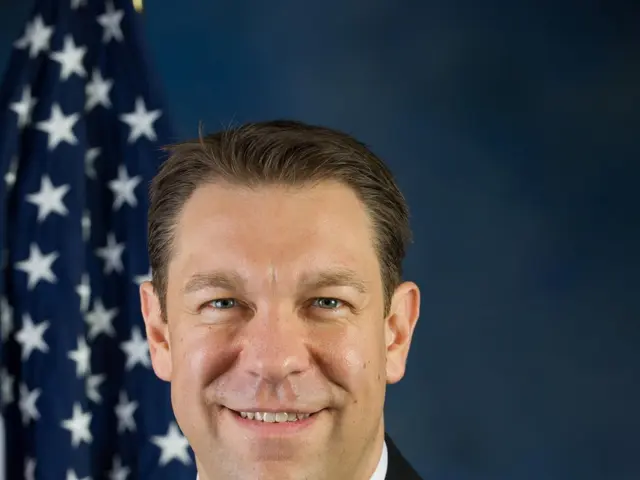 The image shows a man in a suit and tie standing in front of an American flag, with a blue...