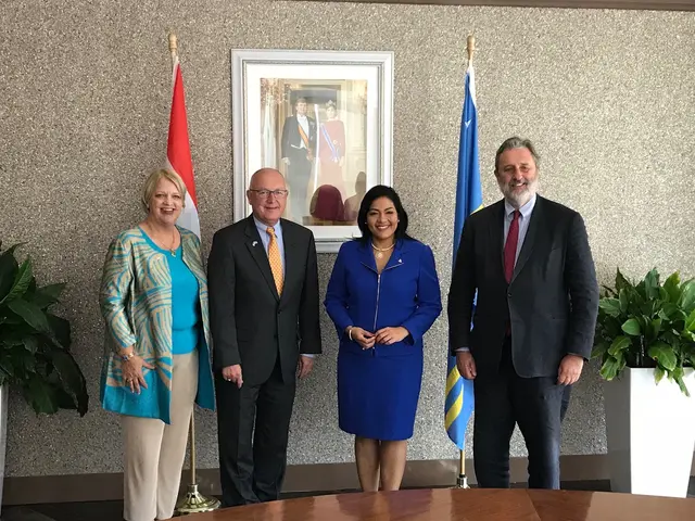 The image shows a group of people standing around a table with papers on it, surrounded by flags...