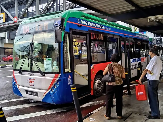 The image shows a blue and white bus stopped at a bus stop with two people standing next to it. The...