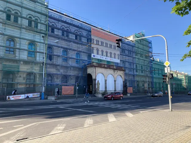 The image shows a large building with a lot of windows on the side of it, surrounded by vehicles on...