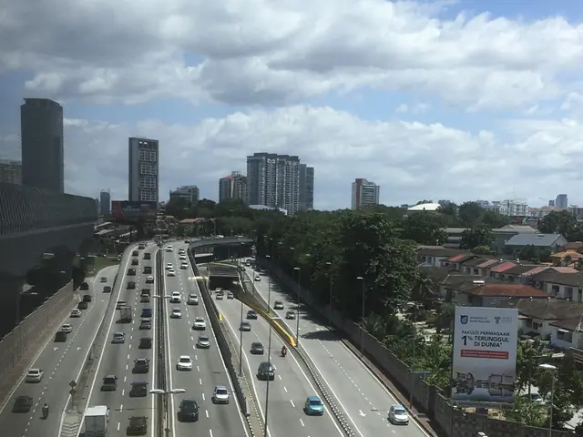 The image shows a highway filled with lots of traffic next to tall buildings, with a bridge on the...