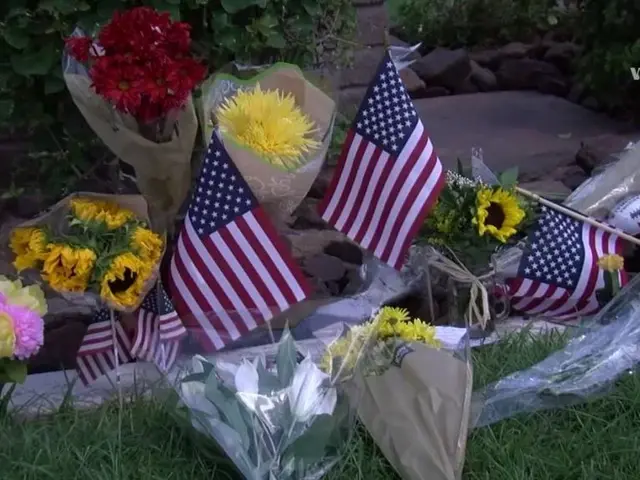 The image shows a solemn scene of flowers and American flags placed on the ground in front of a...