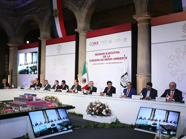 The image shows a group of people sitting at a long table in front of a large screen, surrounded by...