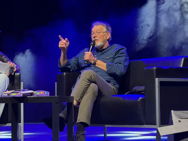 The image shows two men sitting in chairs on a stage, with one of them holding a microphone. On the...