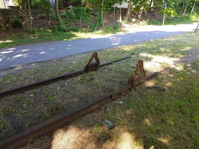 The image shows a train track sitting on the side of a road, surrounded by grass and trees. In the...