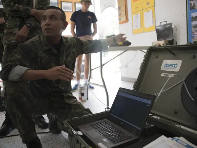The image shows a man in military uniform sitting on the floor next to a laptop computer,...