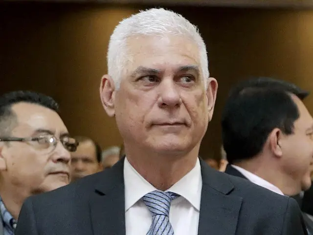 The image shows a man in a suit and tie standing in front of a group of people, with a wall in the...