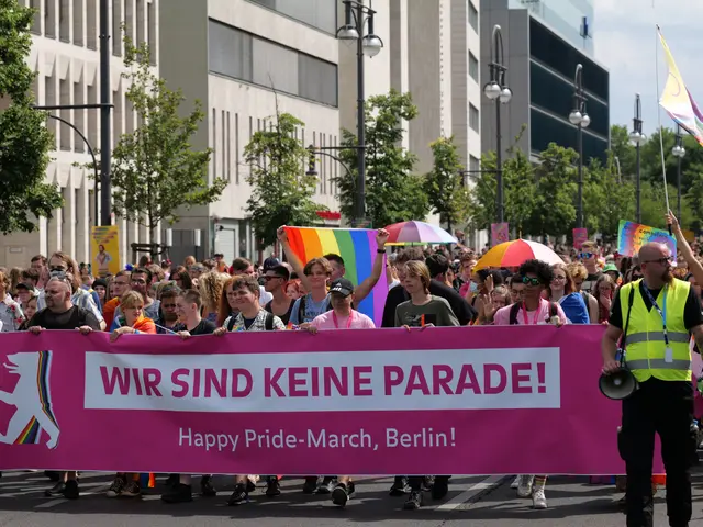The image shows a group of people walking down a street, holding a pink banner with the words...