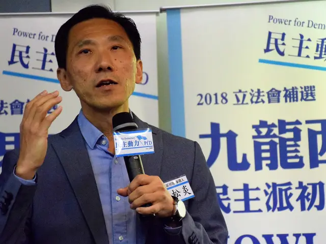 The image shows a man in a suit and tie speaking into a microphone, with banners in the background...