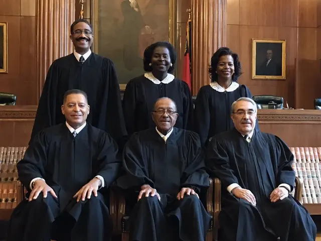 The image shows a group of people sitting in chairs in a courtroom, wearing black robes and...