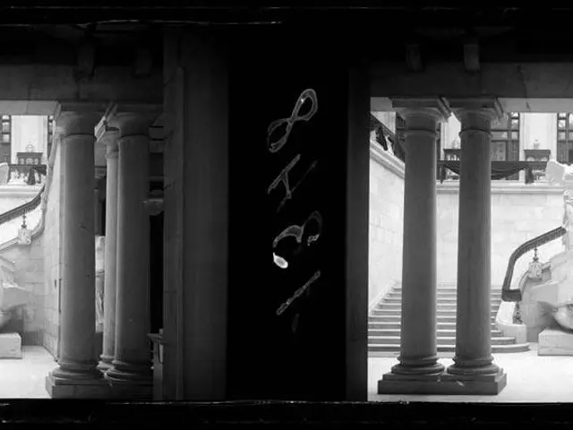 The image shows a black and white photo of a building with columns, statues, stairs, railings, and...