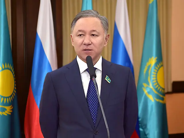 The image shows a man in a suit and tie standing at a podium with a microphone in front of him,...