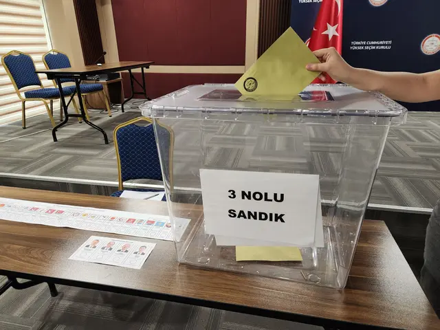The image shows a person putting a voting paper into a ballot box on a table surrounded by chairs,...