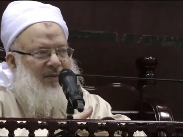 The image shows a man with a white beard and a white turban speaking into a microphone in front of...