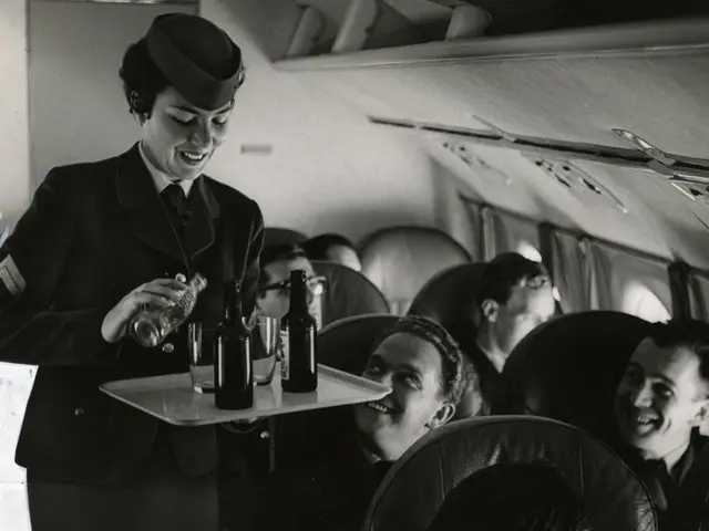 The image shows a black and white scene of a flight attendant serving drinks to a group of...