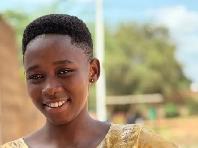 The image shows a young girl in a yellow dress holding a blue phone, with a wall, pole, trees, and...