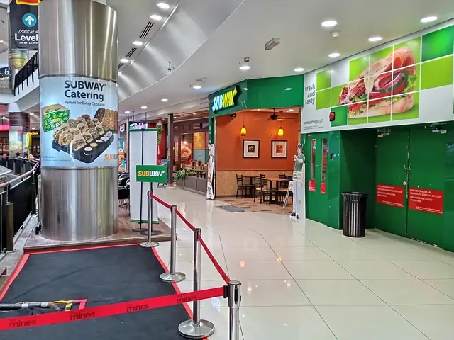The image shows the inside of a subway restaurant with a red carpeted floor, barrier poles with...