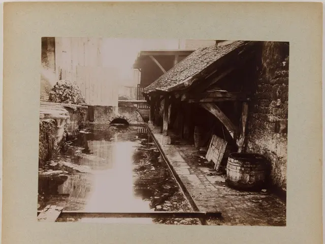 The image shows an old black and white photo of a canal with a building on the right side, a barrel...