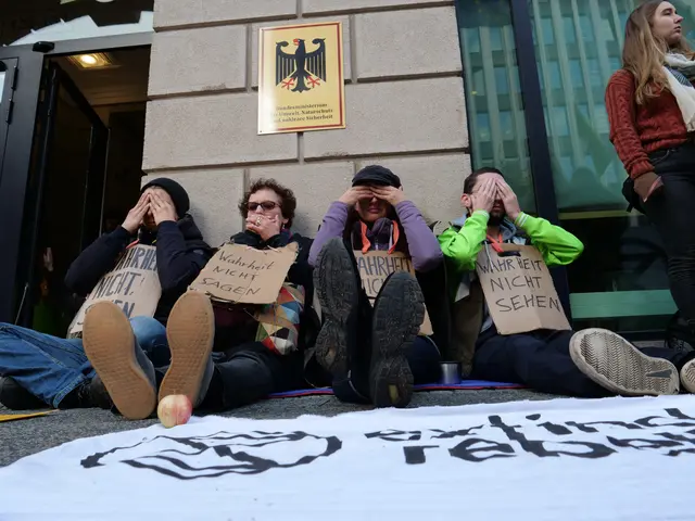 The image shows a group of people sitting on the ground in front of a building, holding signs with...