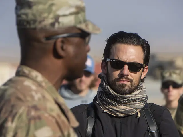 The image shows a man in a military uniform talking to another man wearing a scarf and goggles. The...