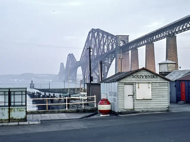 The image shows the Forth Rail Bridge in Scotland, with a road in the foreground, sheds, poles, and...