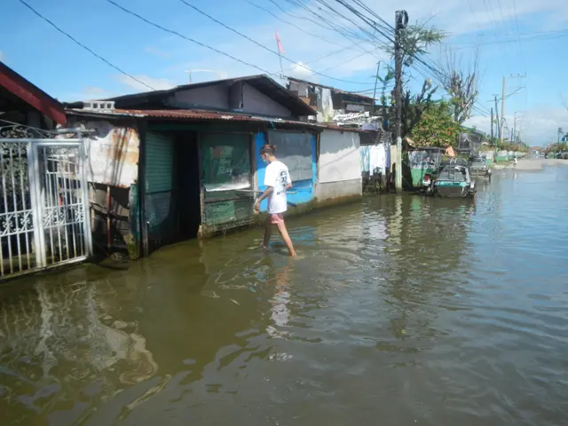 The image shows a man wading through a flooded street in Manila, Philippines. He is wearing a white...