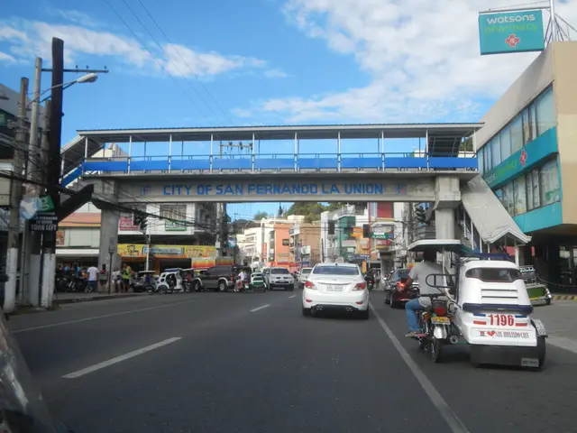 The image shows a bustling city street in San Fernando La Union, with vehicles driving on the road,...