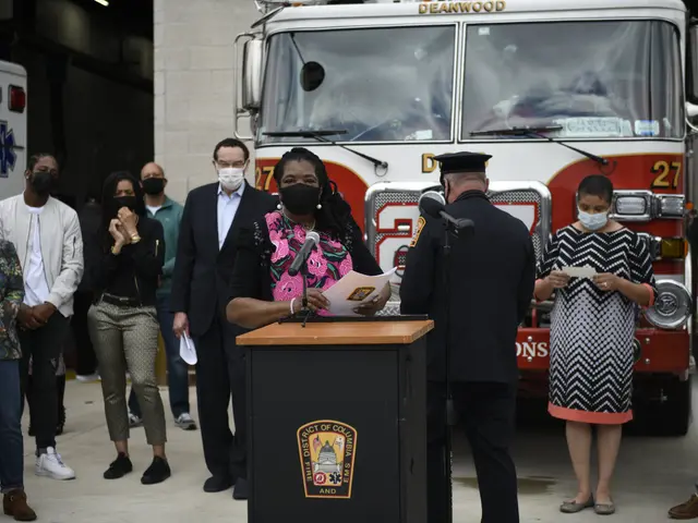 The image shows a woman standing at a podium in front of a fire truck, surrounded by a group of...