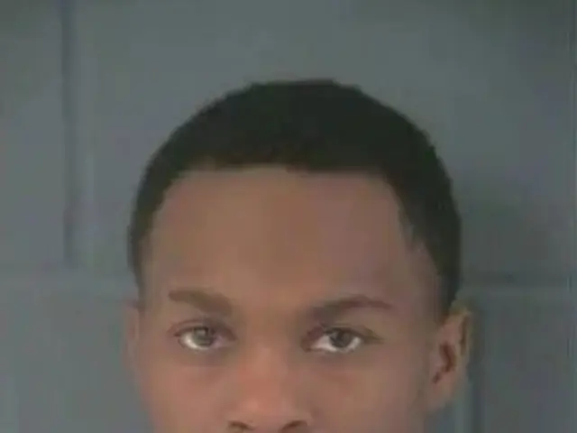 The image shows a mug shot of a man in a white t-shirt, with a wall in the background. The man is...