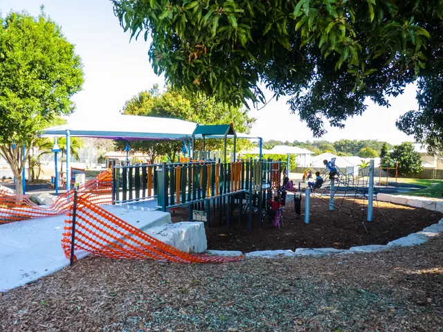 The image shows a park with a playground in the middle of it, surrounded by trees, houses, and a...
