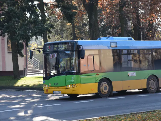 The image shows a green and yellow bus driving down a street lined with trees and a building with...